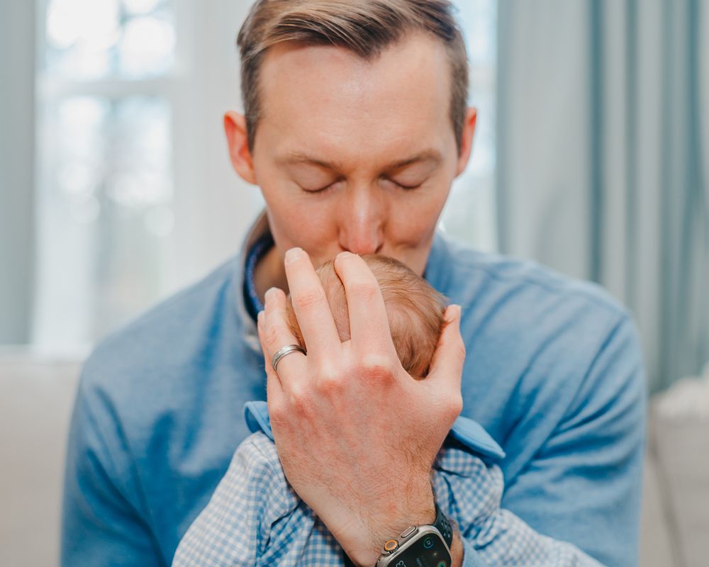 Person wearing a light blue shirt and watch expressing brings their newborn baby to their face to kiss it
