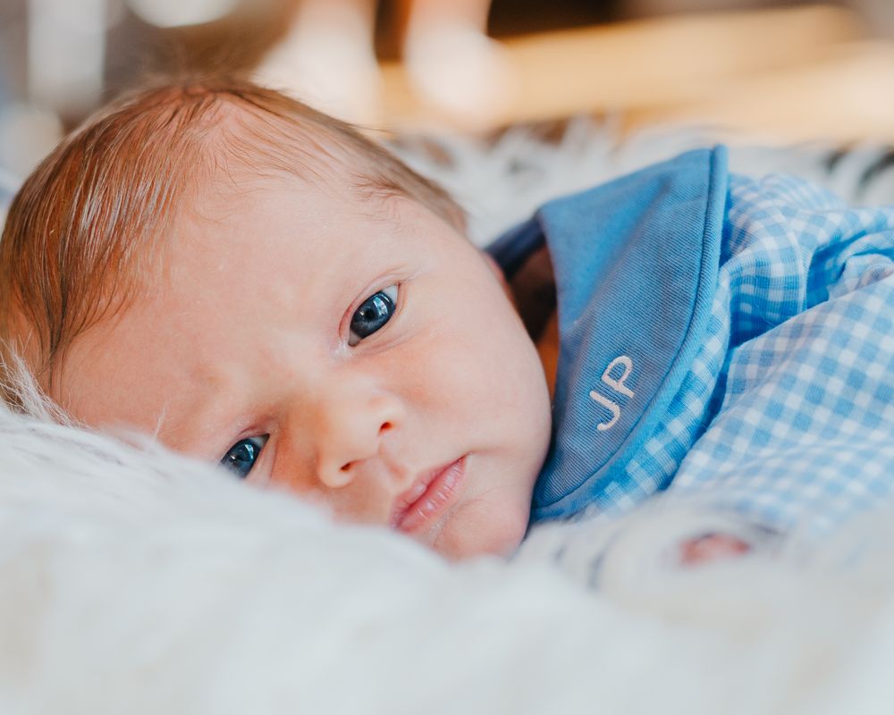 Baby wearing blue bib rests on white bedding while looking at camera with bright blue eyes