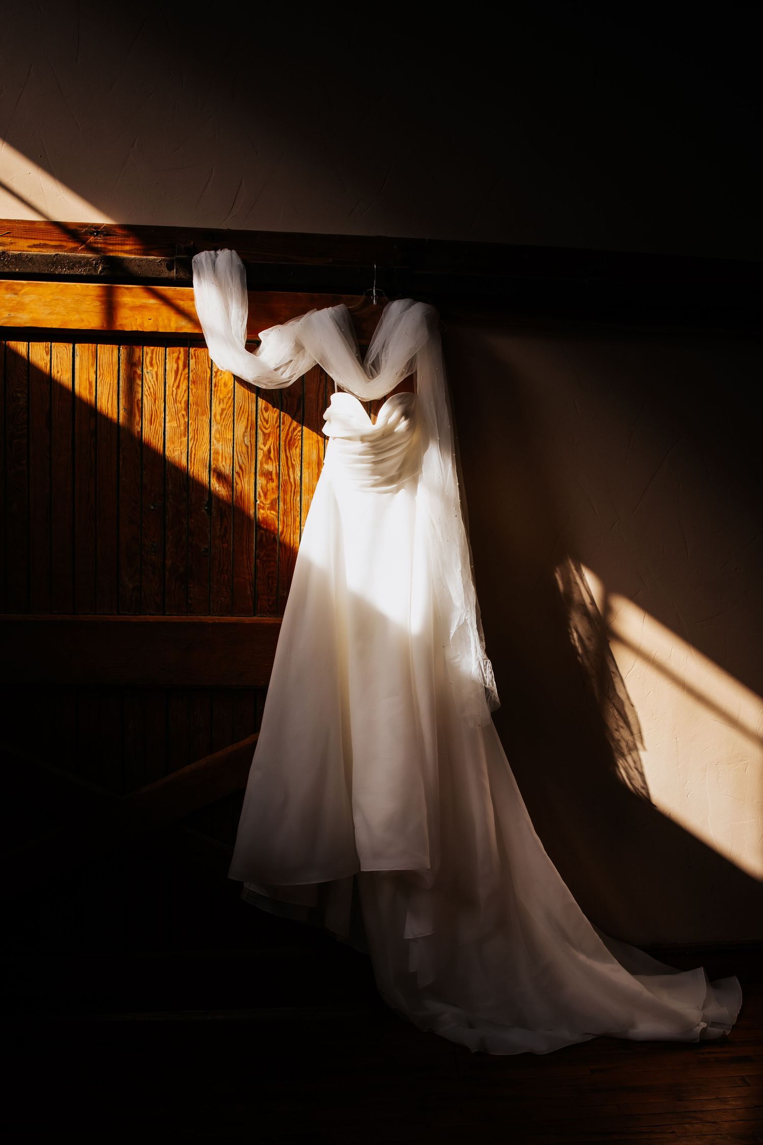 White wedding dress hanging on wooden railing bathed in warm natural light creating dramatic shadows.