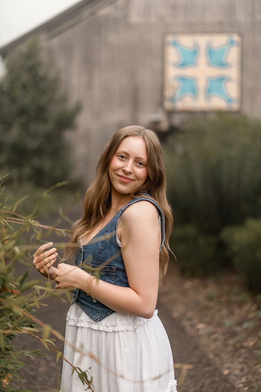 A person wearing a denim vest and white dress poses in front of foliage in an outdoor portrait session.