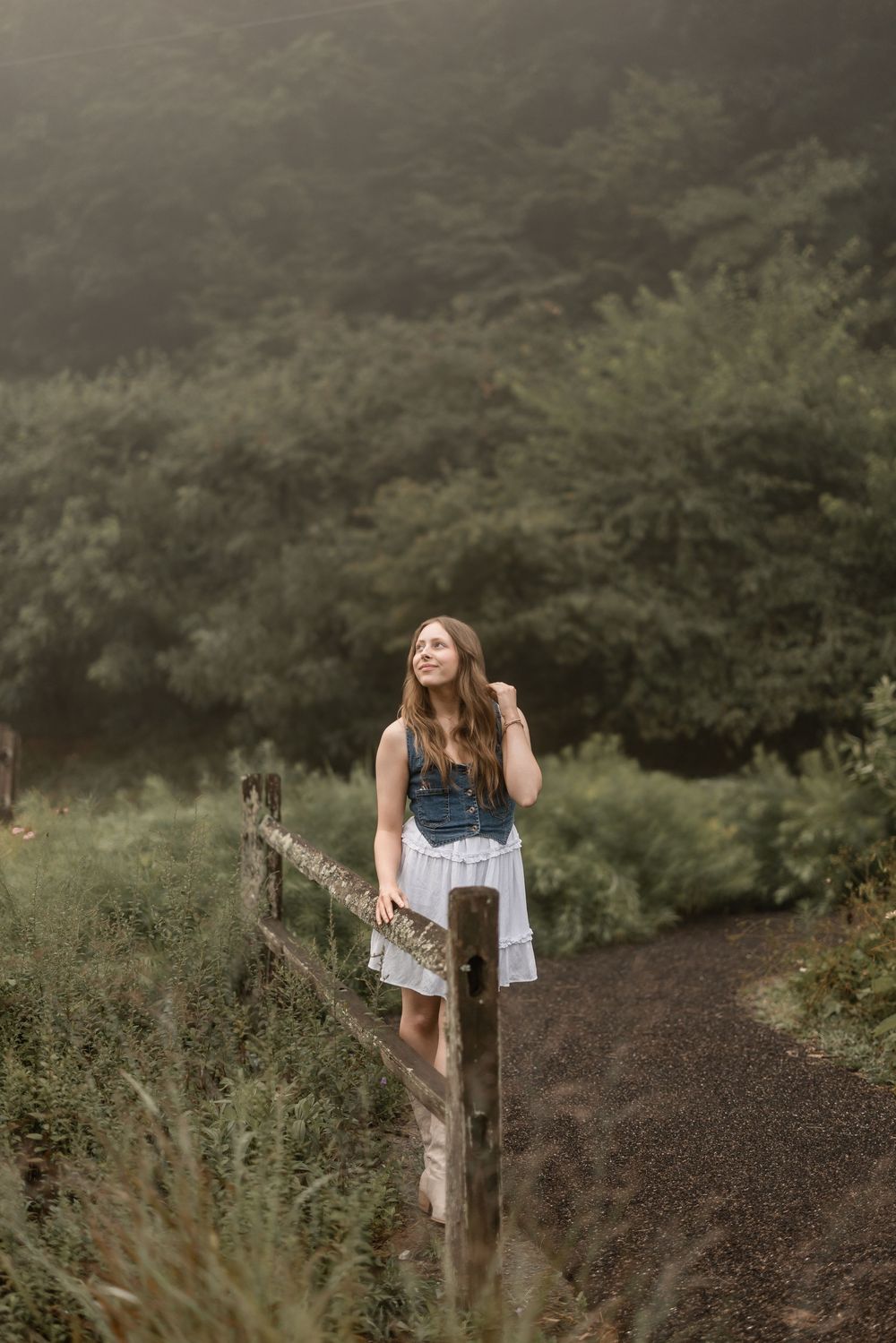 Someone in a denim dress leans against a wooden fence post on a misty forest trail, looking upward dreamily.