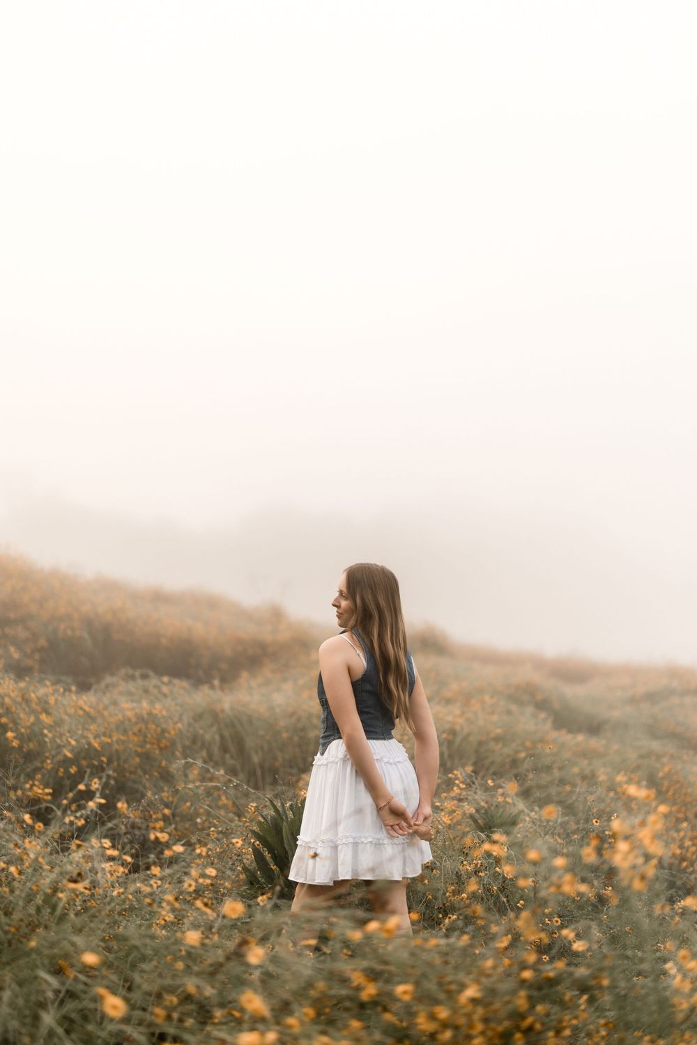 Woman in white dress standing in field of wildflowers on misty hillside during golden hour.