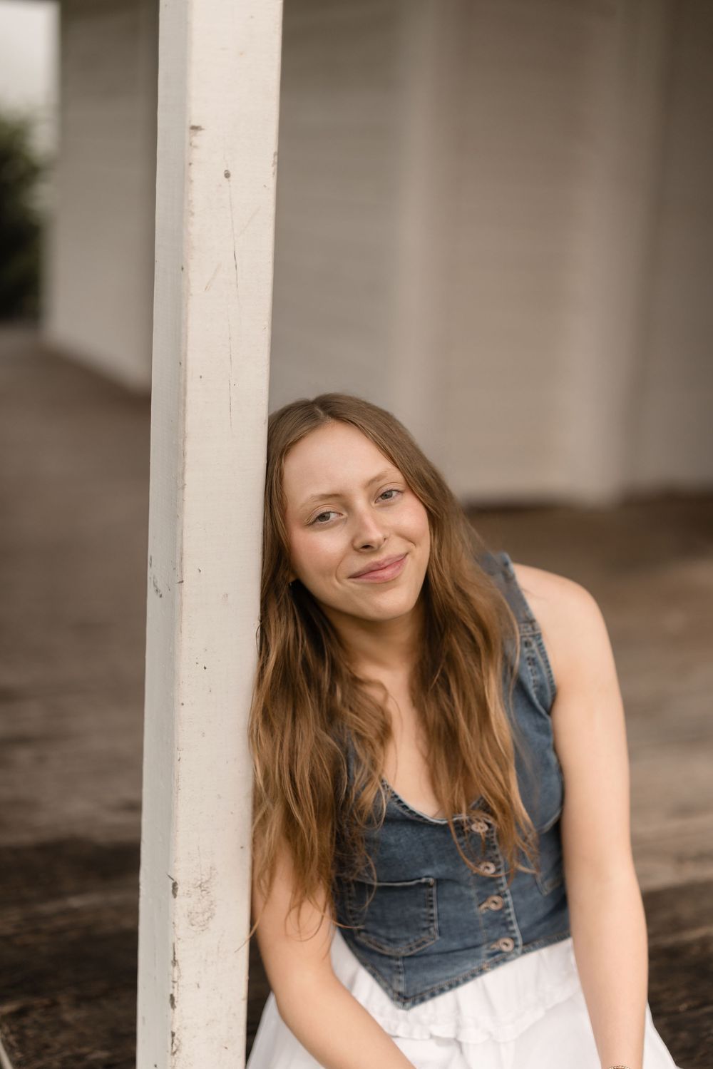 A person in a denim vest leans against a white column on a porch while smiling warmly on a sunny day.