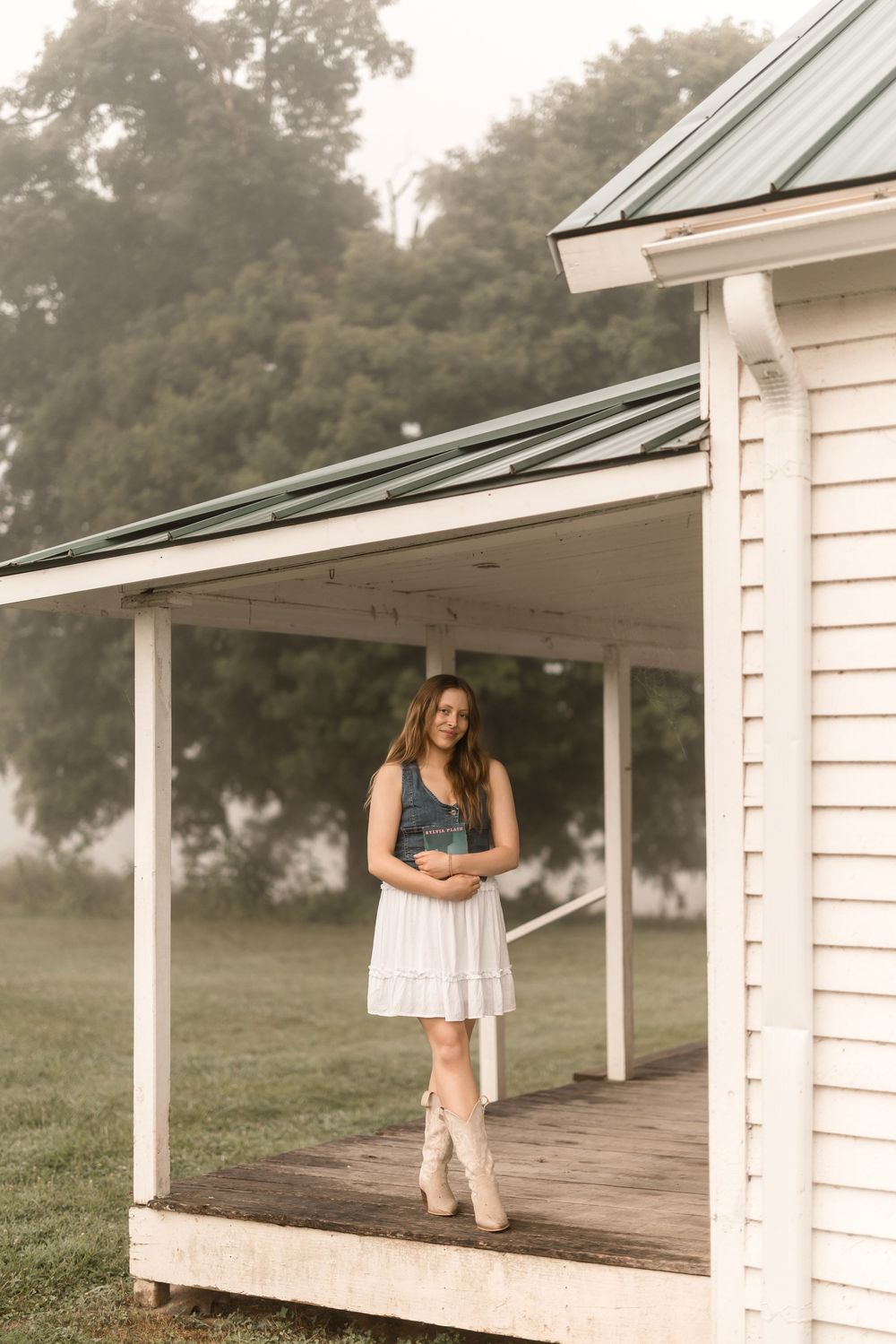Person stands on a rustic farmhouse porch wearing a white dress, denim top and cowboy boots in a foggy country setting.