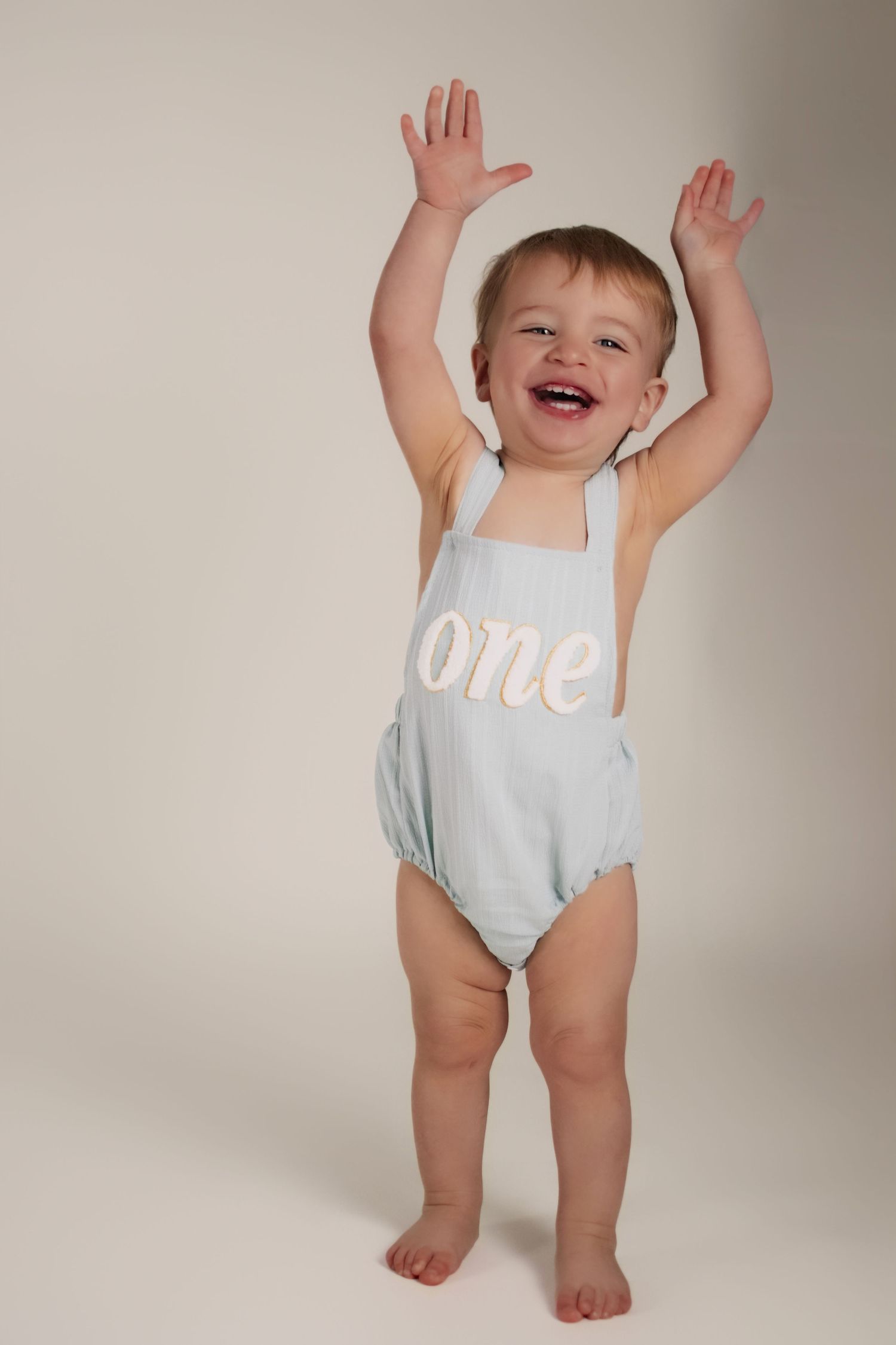 Adorable baby wearing a light blue romper raises their arms playfully in a studio setting.