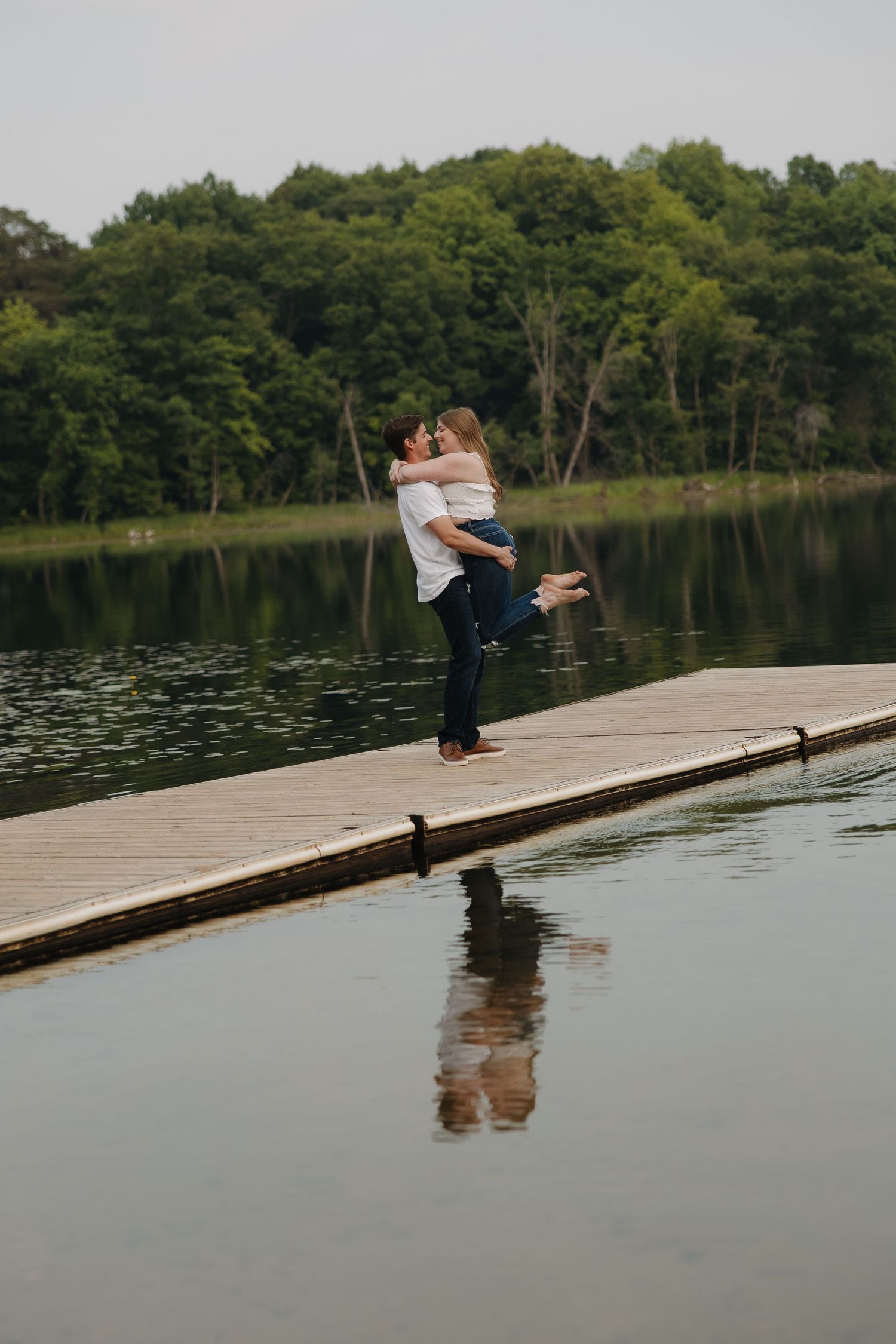 A couple embraces on a wooden dock overlooking a serene lake surrounded by lush green forest.