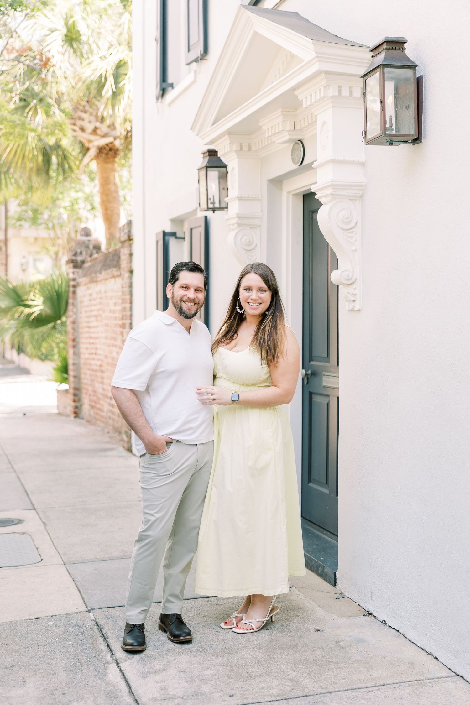 A couple stands in front of a classic Charleston-style home with white walls and green doors flanked by lantern sconces.
