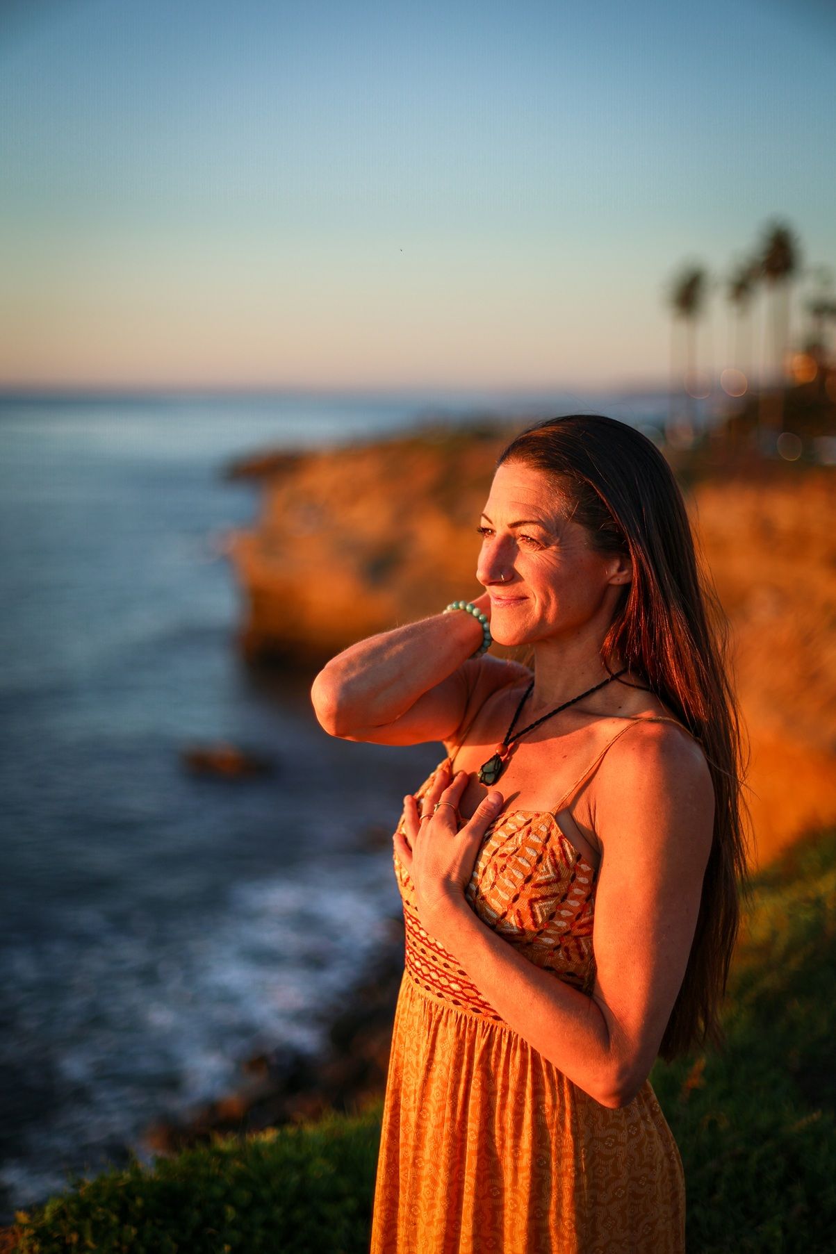 Woman in orange dress with hands on chest enjoying golden sunset by coastal cliffs.