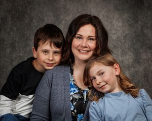 Smiling woman in a gray cardigan poses for a studio family portrait with a young boy and a girl leaning in against a textured gray backdrop.