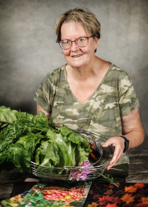 Smiling woman with glasses in camo shirt holding a wire basket of fresh garden vegetables at a wooden table.