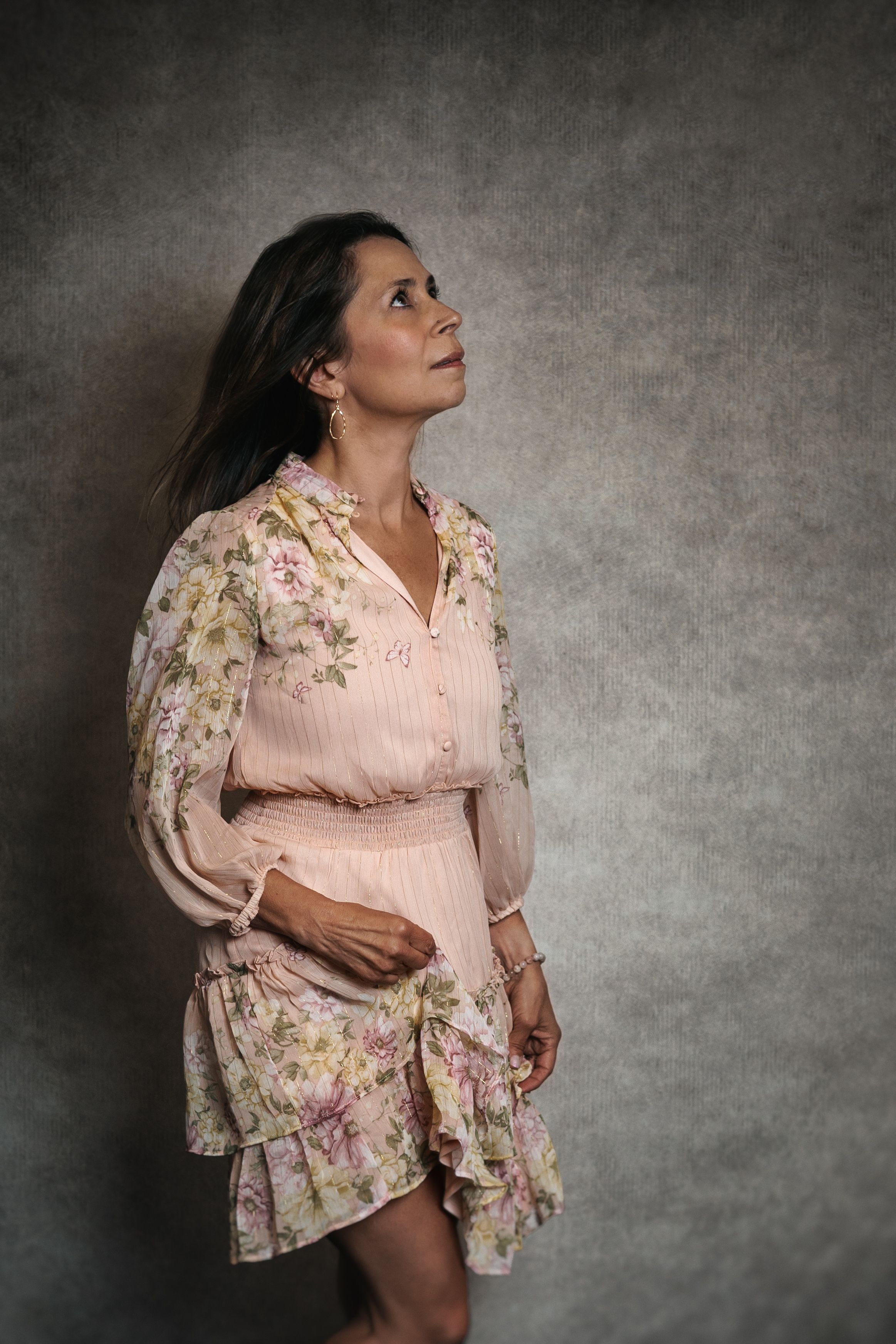A pregnant person in a feminine pink floral dress poses against a dark textured backdrop in a maternity photoshoot.