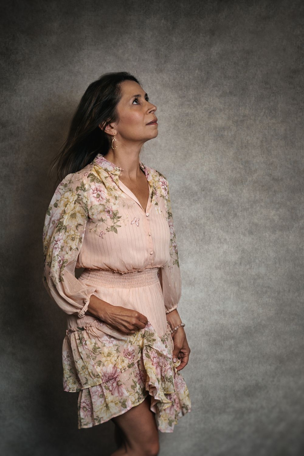 A pregnant person in a feminine pink floral dress poses against a dark textured backdrop in a maternity photoshoot.