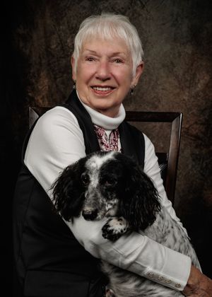 Smiling older woman with short white hair in a black vest and white turtleneck holds a black and white spaniel in a studio portrait.