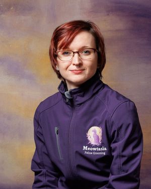 Woman with short red hair and glasses smiling in a purple Meowtasia Feline Grooming jacket against a painted backdrop.