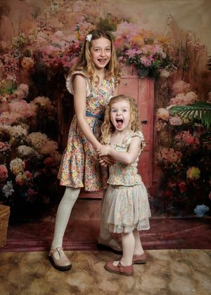 Two children in floral dresses pose against a romantic pink floral backdrop in a studio photography setting.