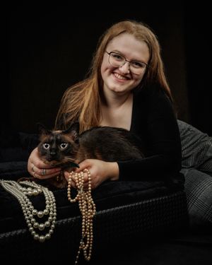 Smiling woman with long red hair and glasses holds a dark Siamese cat while draped with strands of pearl necklaces on a black velvet sofa.