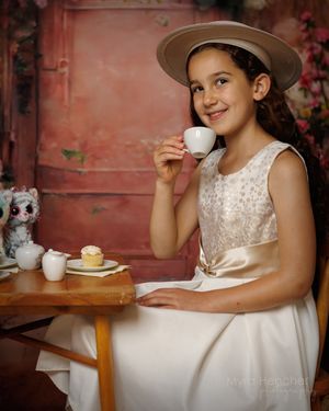 Young girl in white dress and tan hat holds teacup at small table with cupcake and toy at a tea party photo shoot.