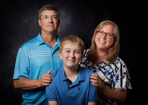 Family portrait of two adults and a young boy smiling, wearing blue, against a dark studio background.