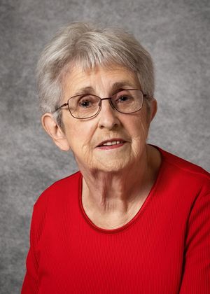 Portrait of an elderly woman with short gray hair and wire-rimmed glasses, smiling softly in a red top against a gray studio background.