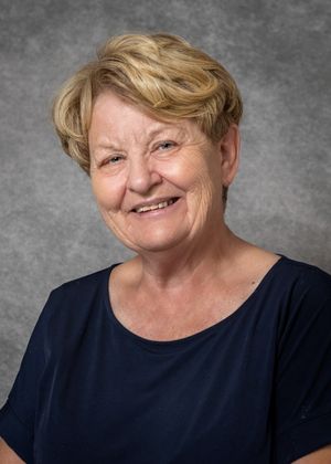 Smiling older woman with short blonde hair wearing a navy blue top, professional headshot on gray background.