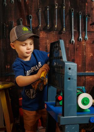 Young boy in John Deere cap and Caterpillar shirt playing with toy workbench tools in a garage workshop.