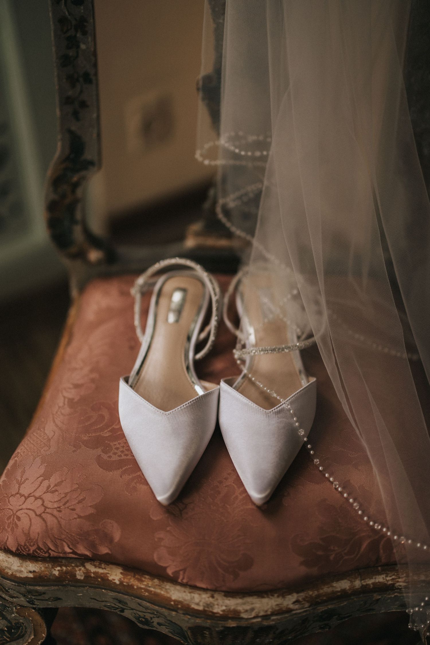 Elegant white bridal shoes with pointed toes photographed on a rustic brown chair through a delicate wedding veil.