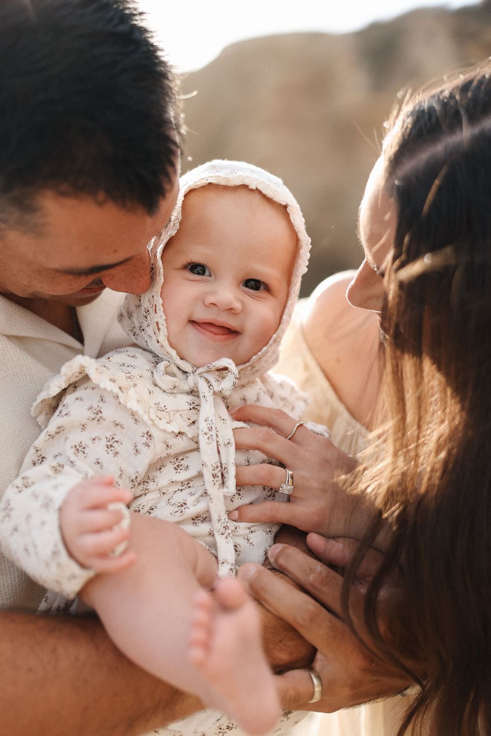Sweet family moment showcasing precious baby wearing white lace bonnet and dress during golden hour photoshoot outdoors.