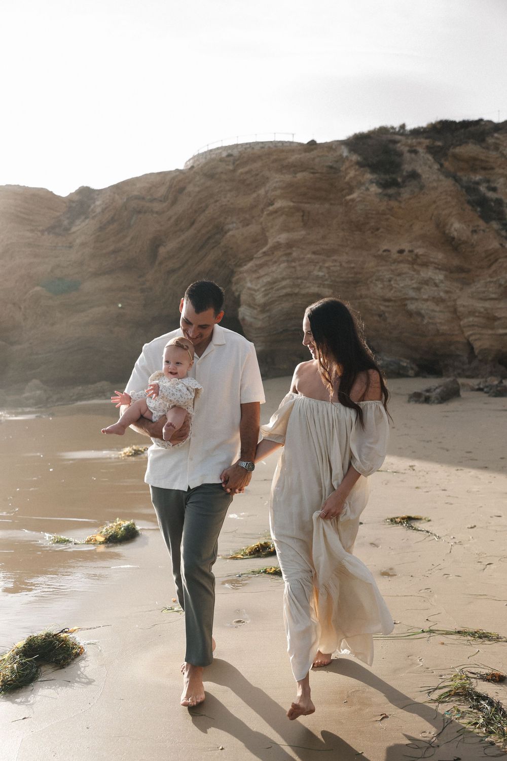 Young couple in white and grey attire share tender moments while walking along a scenic beach with cliffside backdrop.