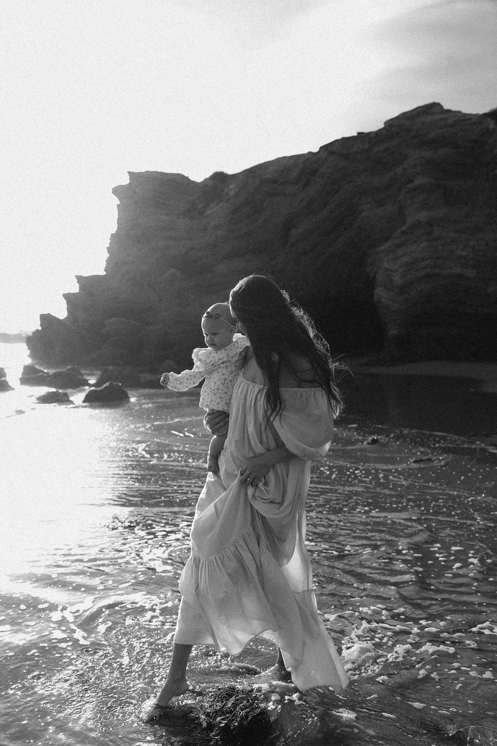 Couple in white dresses dancing and embracing on a rocky beach with dramatic cliffs in black and white photography.