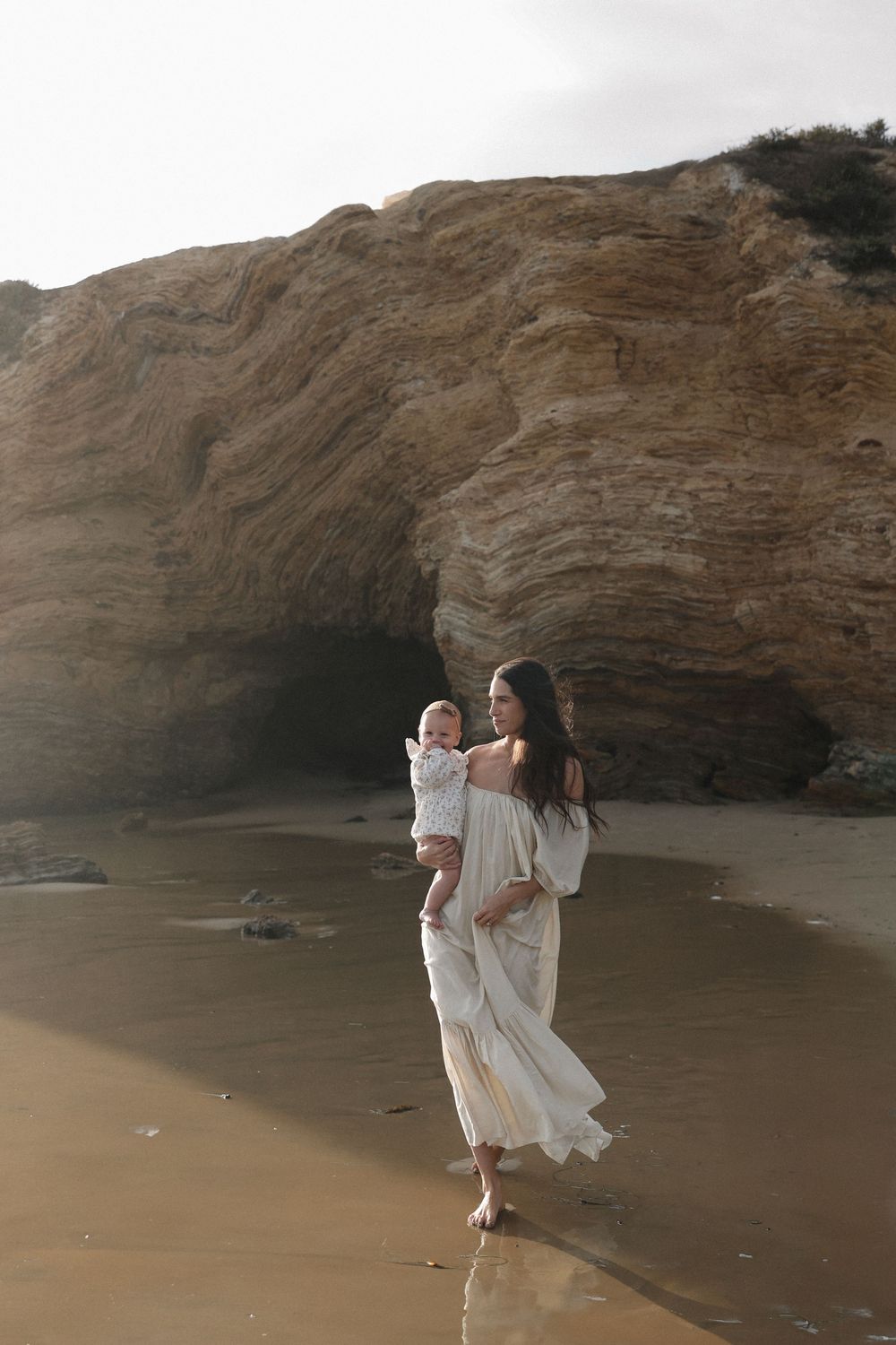Two silhouettes walking along a sandy beach near dramatic coastal cliffs at sunset with soft sunlight filtering through.