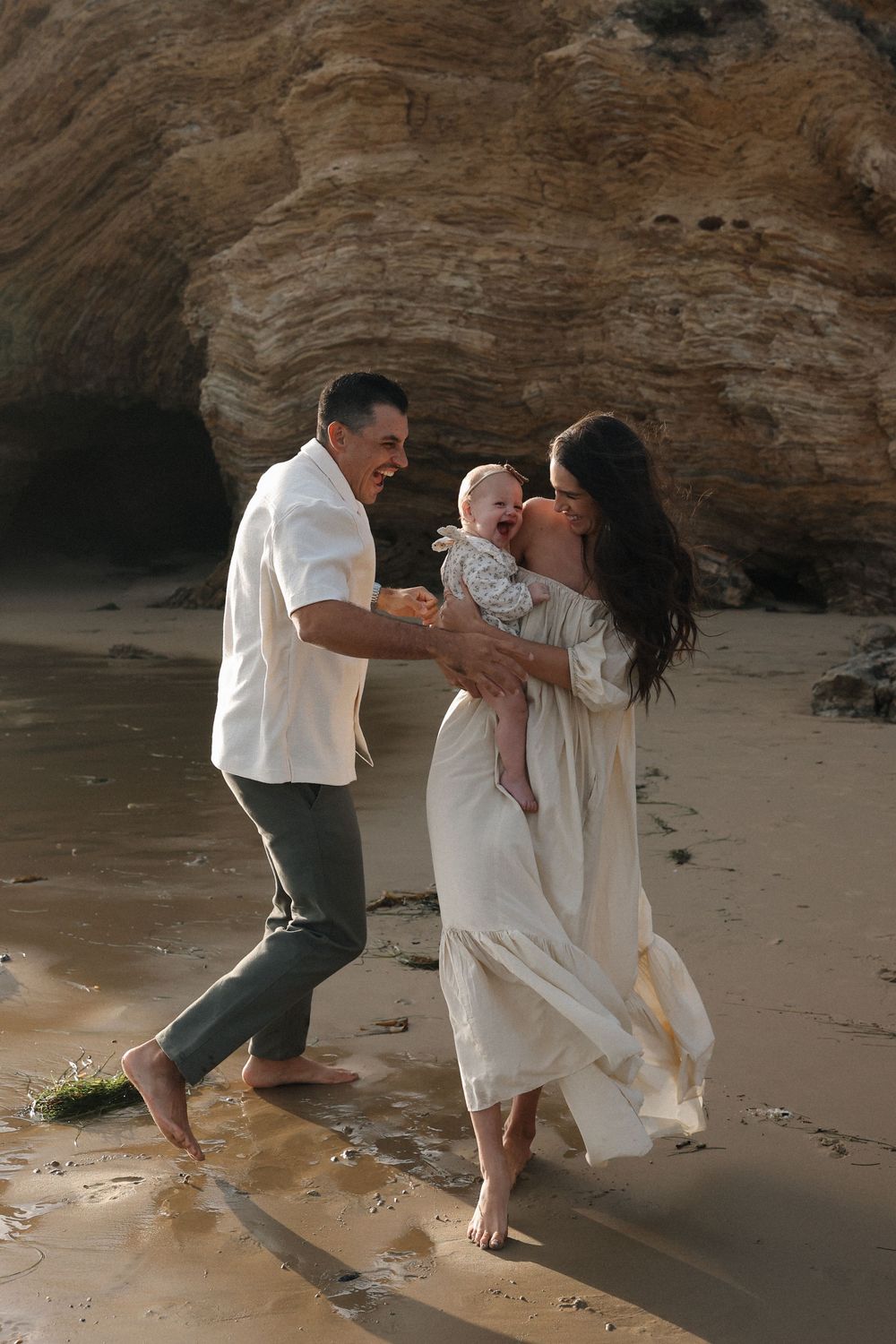 Family enjoying a tender moment together on a beach near rocky cliffs as their white clothing flows in the ocean breeze.