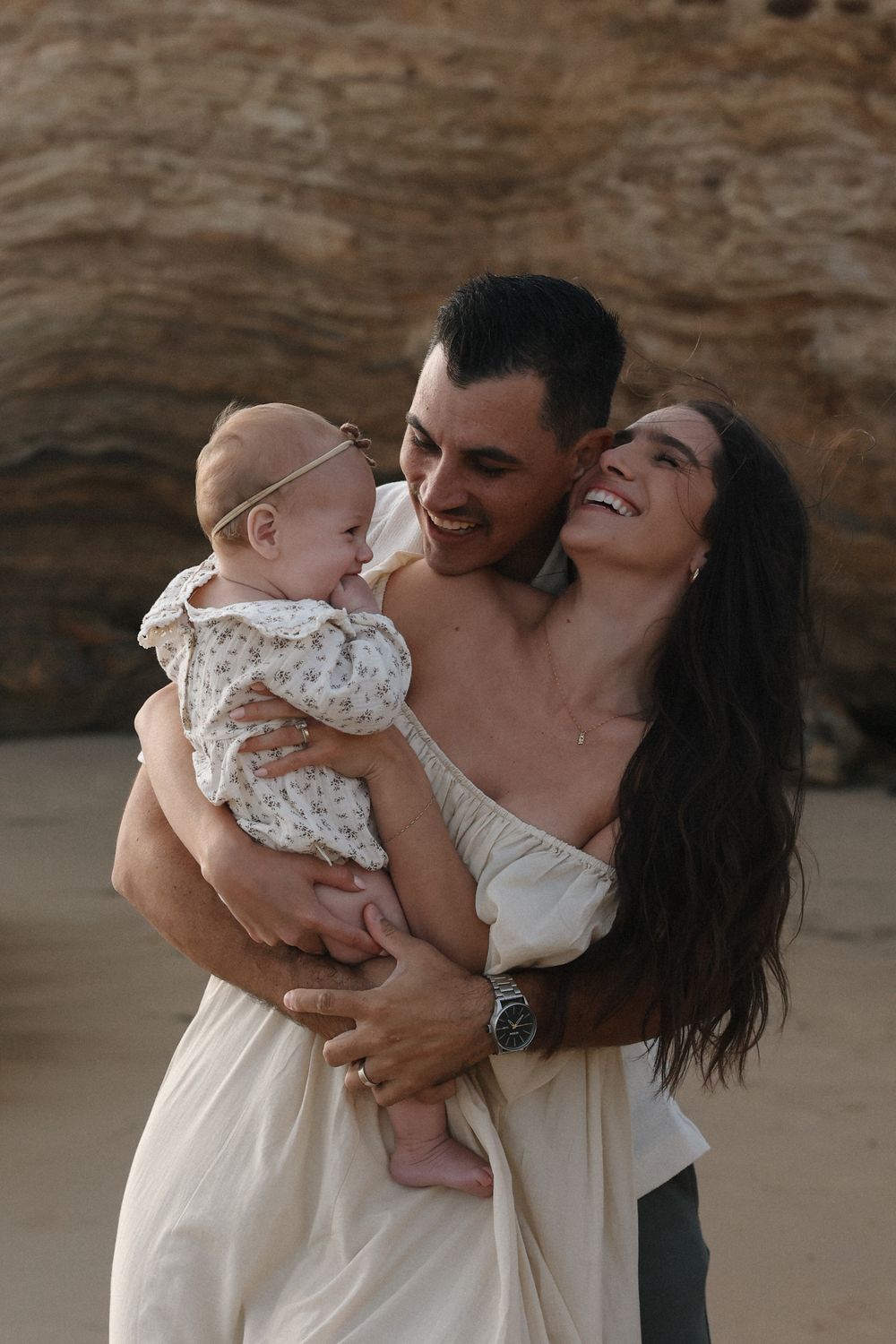 A family shares joyful laughter together on a beach with rocky cliffs behind them.
