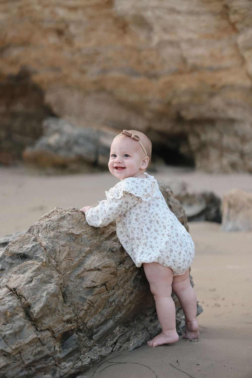 A baby in a floral onesie crawls and plays near large rocky outcrops on a sandy beach.
