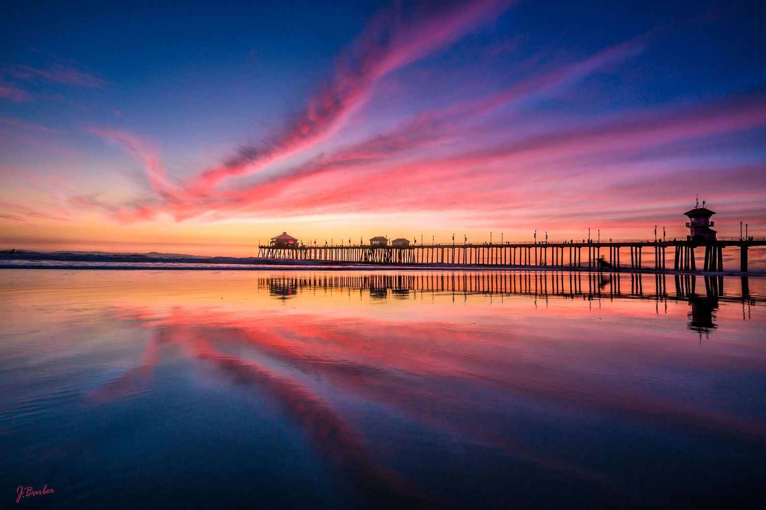 Vibrant red and purple sunset clouds reflect dramatically in still water, creating a mesmerizing mirror-like panorama at hungtington beach.