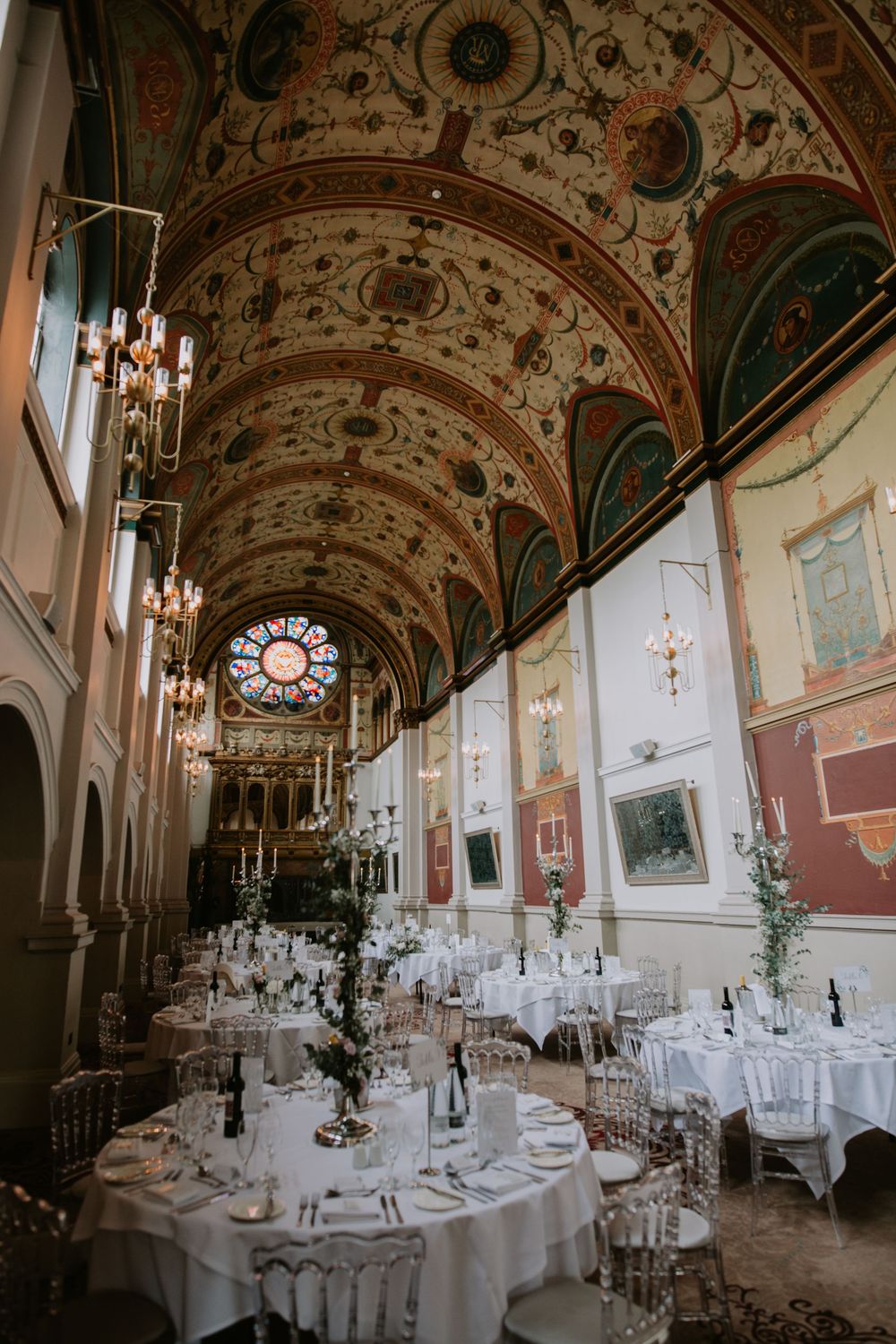 An elegant historic ballroom with ornate ceiling frescoes, crystal chandeliers, and white-clothed round tables for a wedding reception.