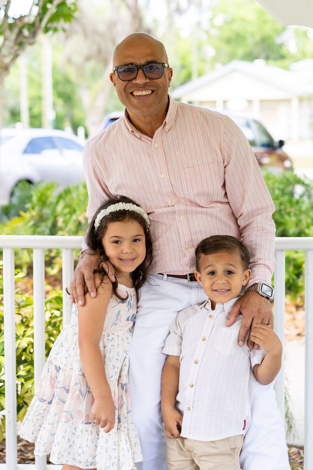 Family portrait in coordinated white and beige outfits under a shaded garden pergola.