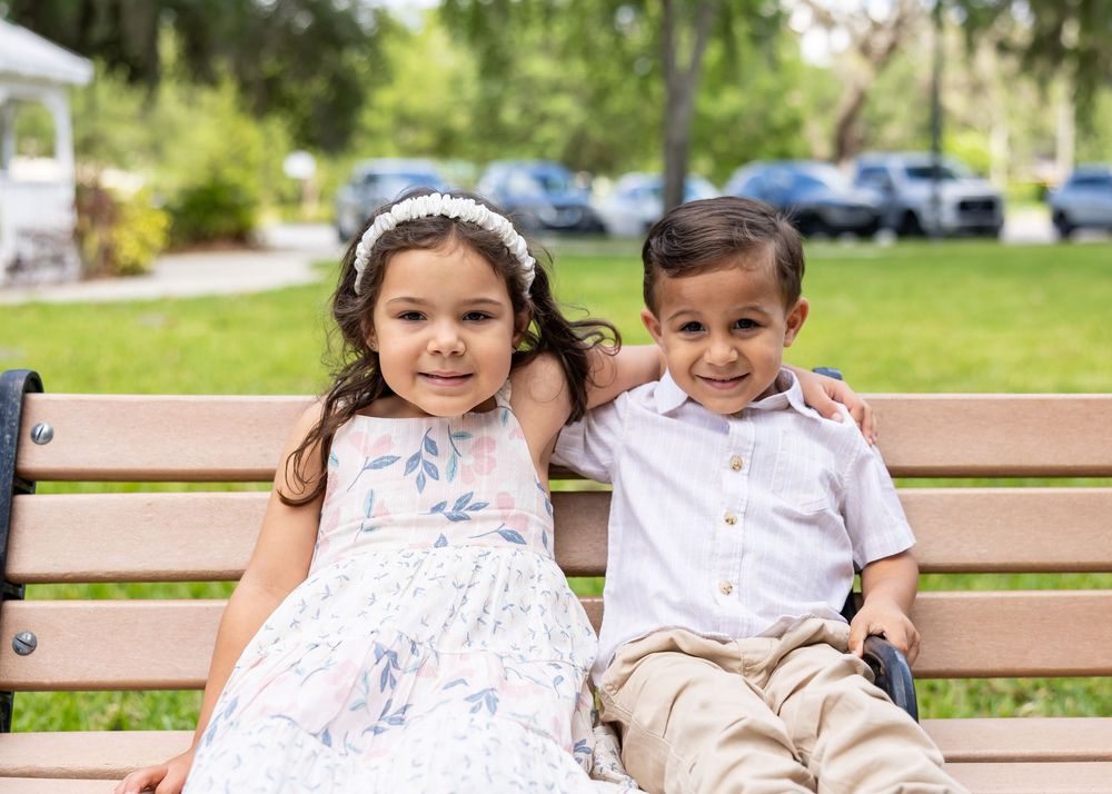 Family members sit together on a wooden park bench in matching light-colored outfits.