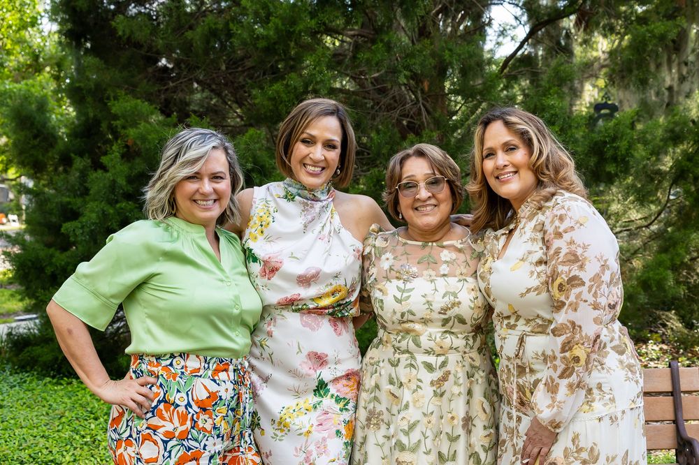 Four people in floral and colorful dresses pose together in a lush garden setting.