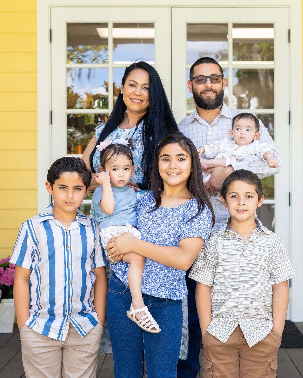 Large family gathering in coordinated blue and neutral outfits posed by yellow house.