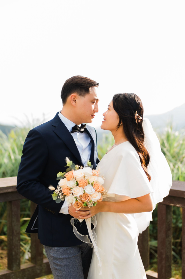 A romantic outdoor wedding photo of a couple in white shirts embracing and laughing among spring blossoms and greenery.