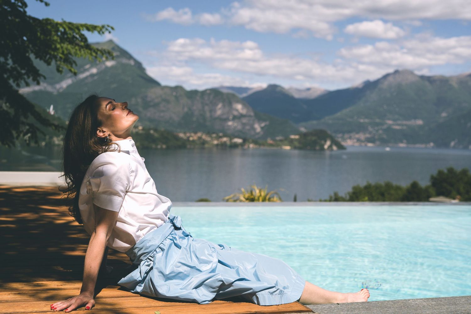 Donna in abiti estivi si rilassa seduta a bordo piscina con vista su Bellagio e il Lago di Como.