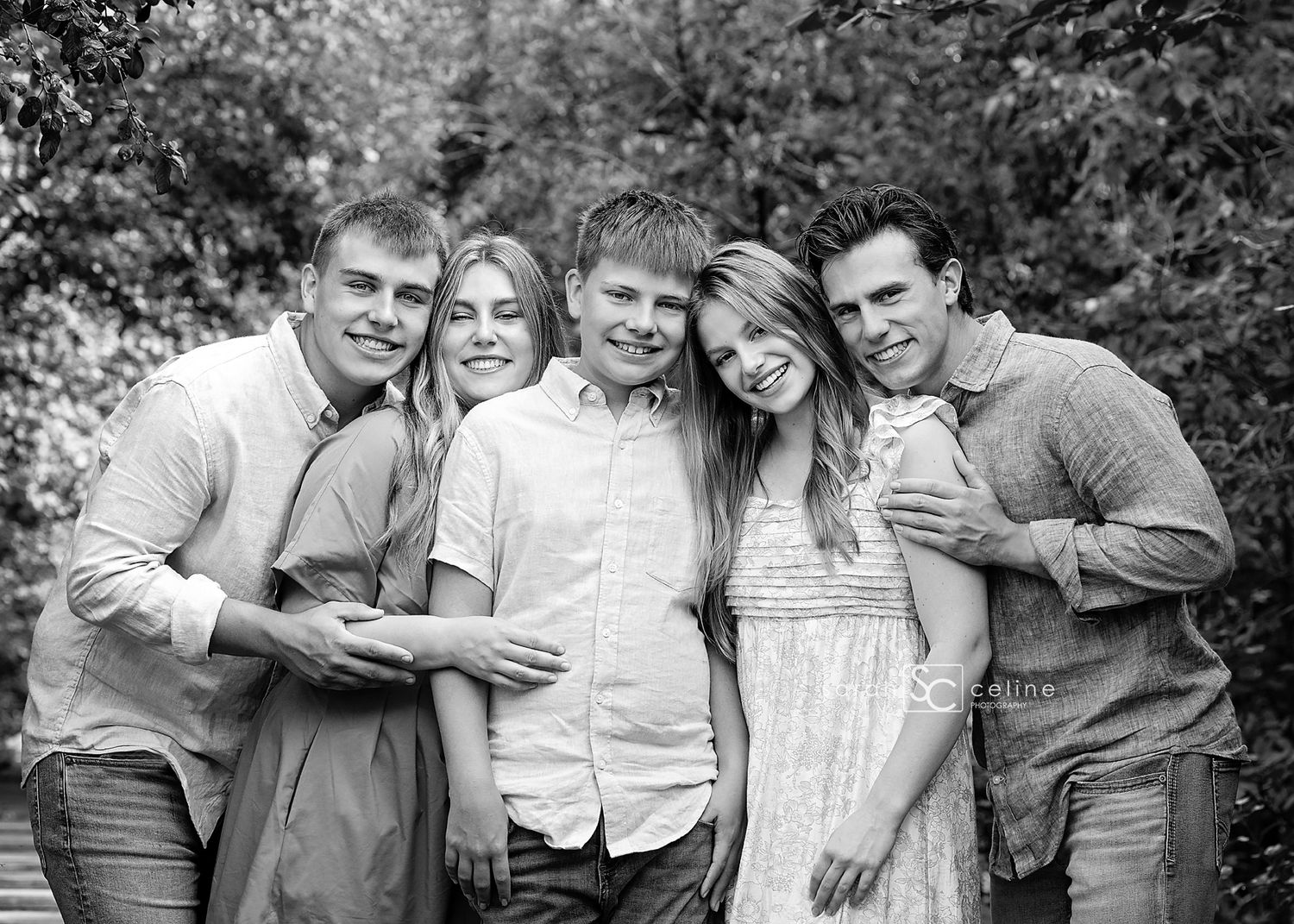 Black and white group portrait outdoors with five people smiling together in a casual setting against trees.