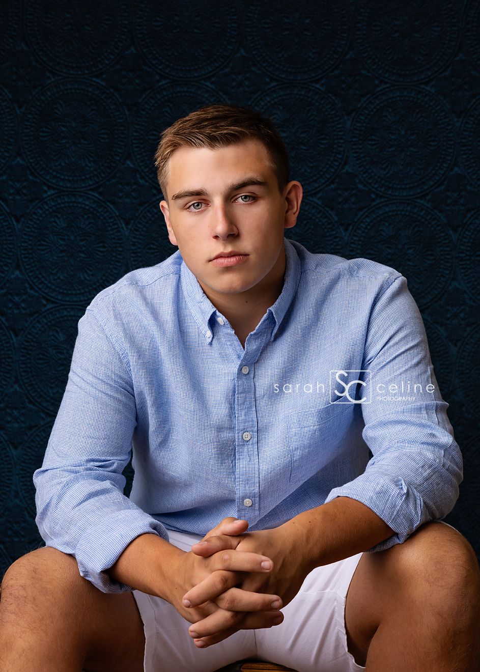 Professional studio portrait of someone wearing a light blue button-down shirt and khaki shorts against a textured navy background.