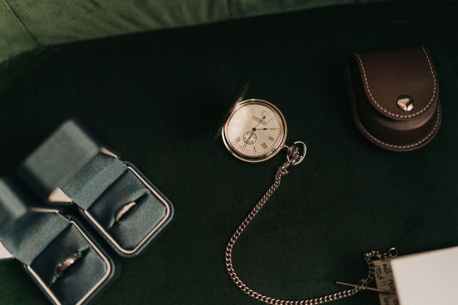 Vintage pocket watch with leather case and wedding rings displayed on dark background in moody lighting.