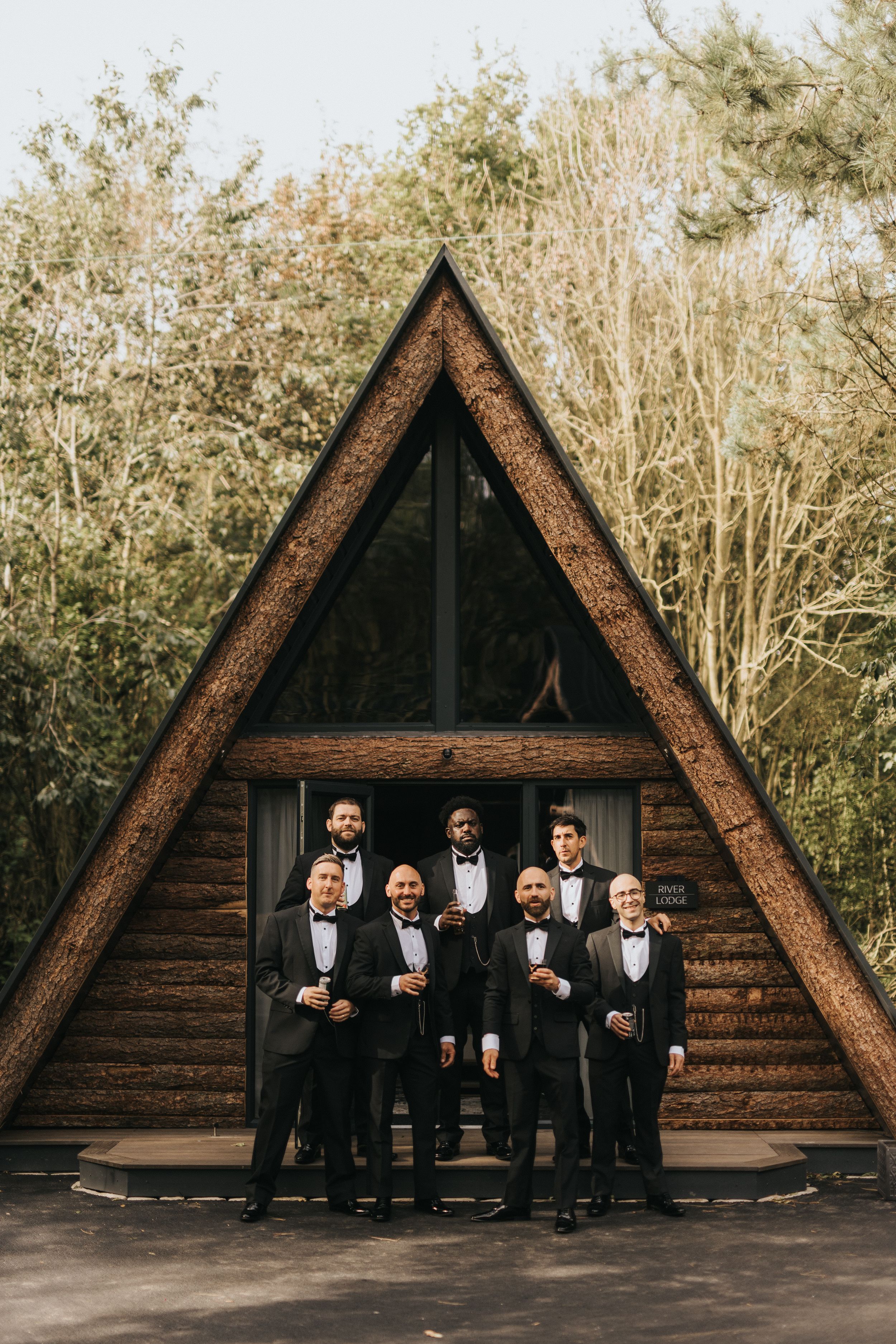 Groomsmen in black tuxedos pose together in front of rustic wooden A-frame cabin at Grand Lodge Oaklands - taken by Grimsby photographer.