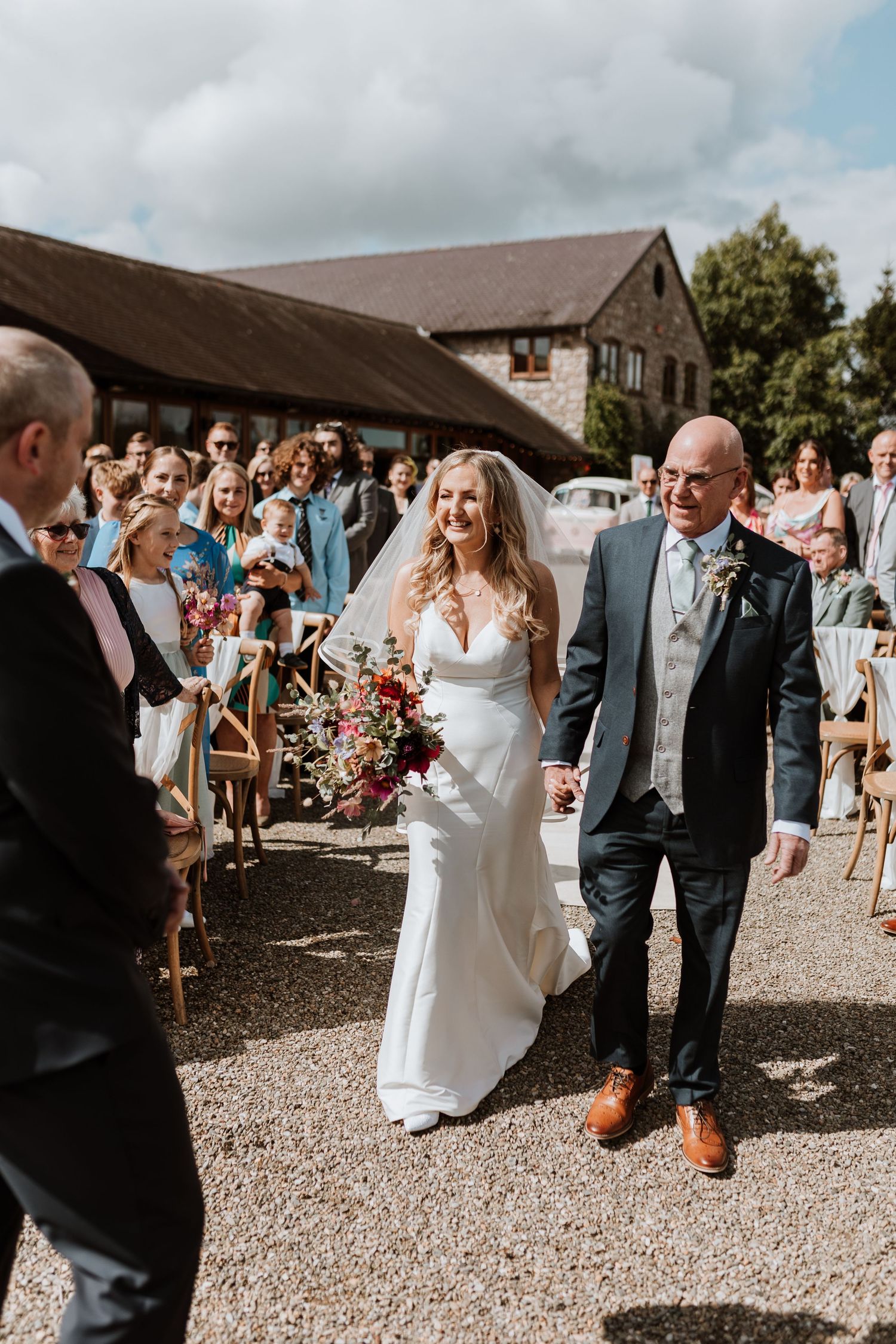 A sunny outdoor wedding ceremony at a rustic barn venue with guests watching the couple walk down the aisle.
