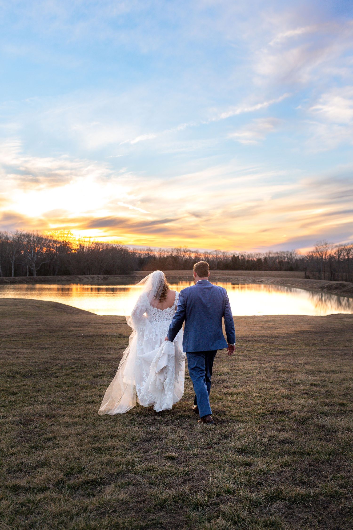 Couple walking along lakeside at sunset with flowing wedding dress trailing behind.