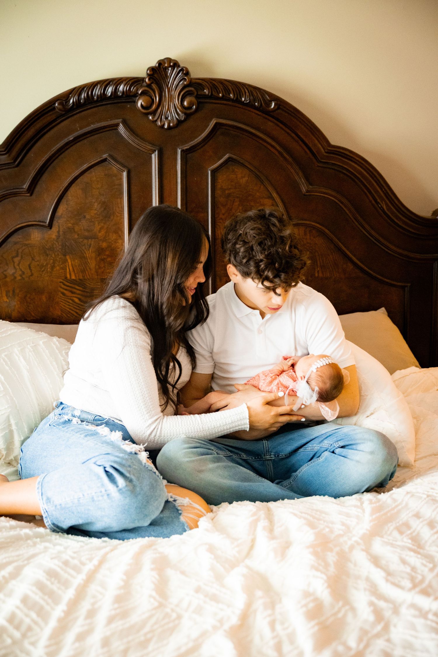 New parents sitting on bed with ornate wooden headboard, tenderly caring for their newborn baby together on white bedding.
