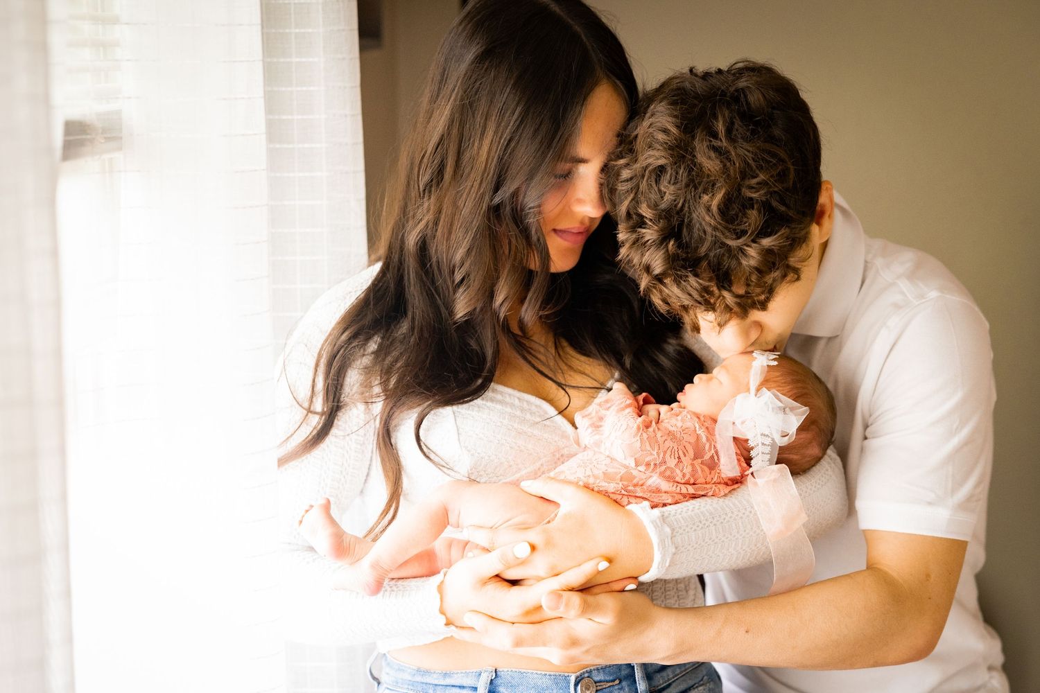 A loving couple tenderly holds their newborn baby wrapped in a soft peach blanket in a warmly lit room.