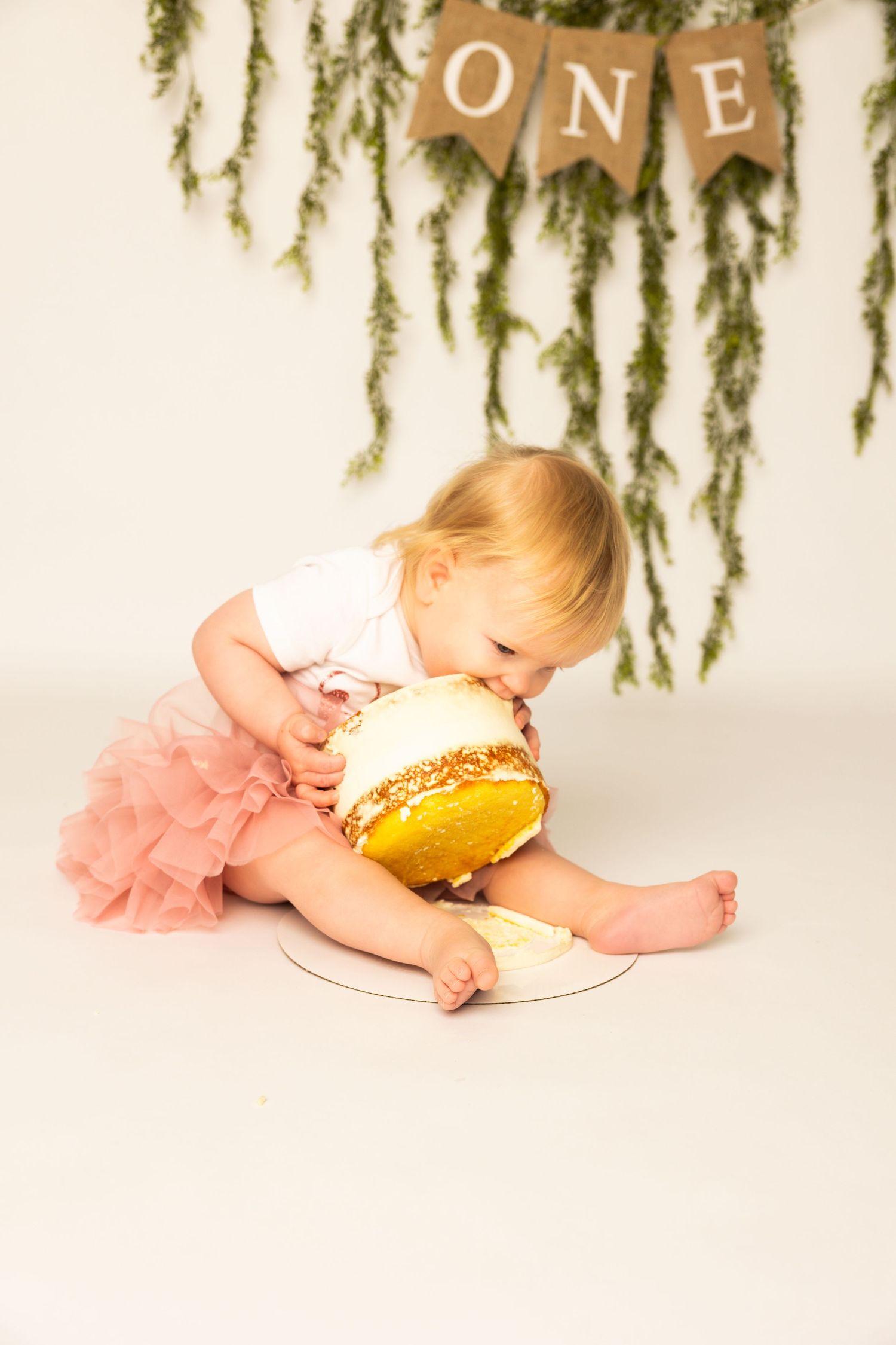 Baby in pink tutu dress smashing and tasting yellow frosted birthday cake during cake smash photo session.
